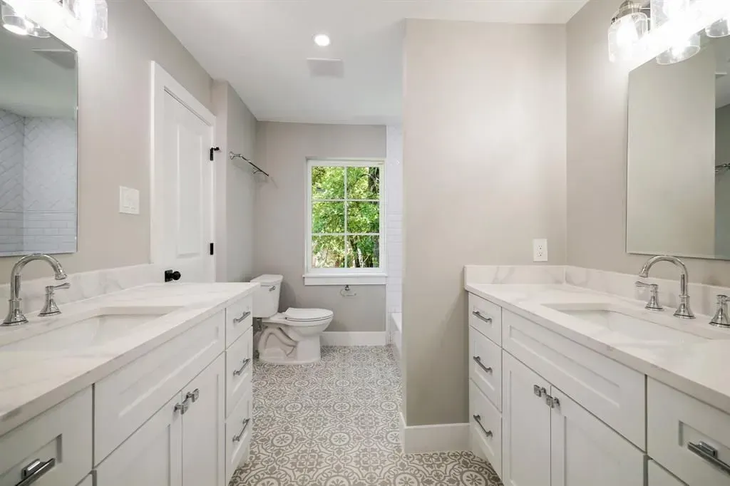 Bright modern bathroom with double sinks, white cabinets, patterned tile floor, and a window view.