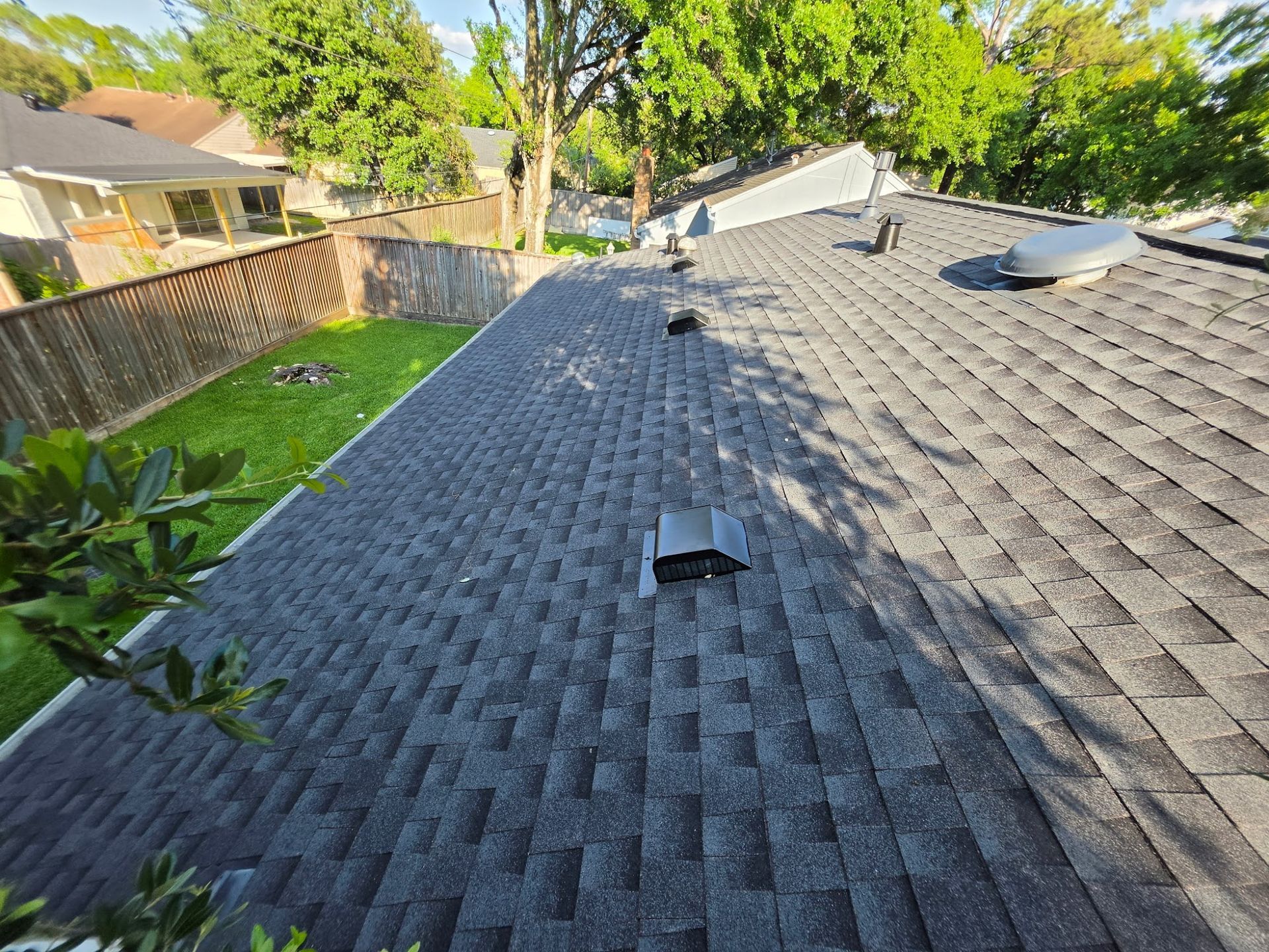 Rooftop with dark gray shingles, skylights, and a vent, beside green trees and a fenced yard