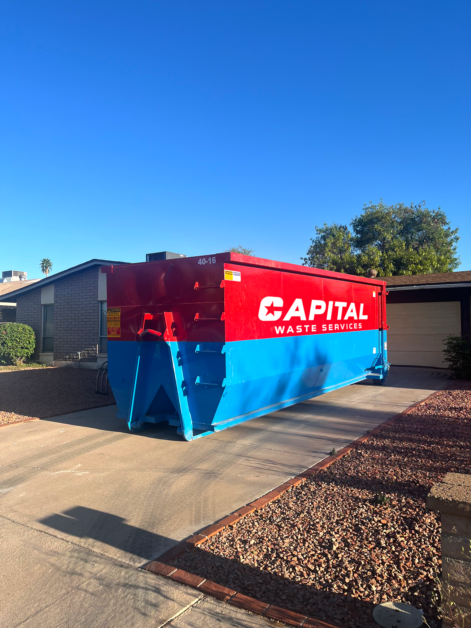 A large red and blue Capital dumpster sits on a residential driveway in front of a house under a sunny blue sky.