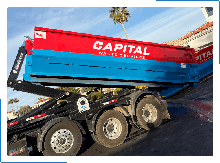 A red and blue Capital Waste Services dump truck trailer being loaded onto a transport truck in a sunny outdoor setting.