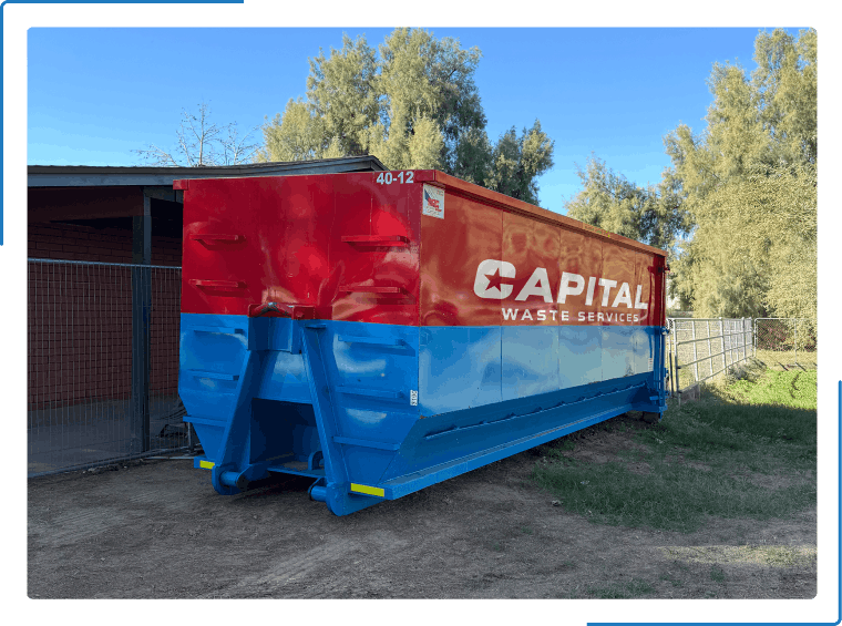 A red and blue Capital Waste Services roll-off dumpster sitting on a dirt lot outdoors.