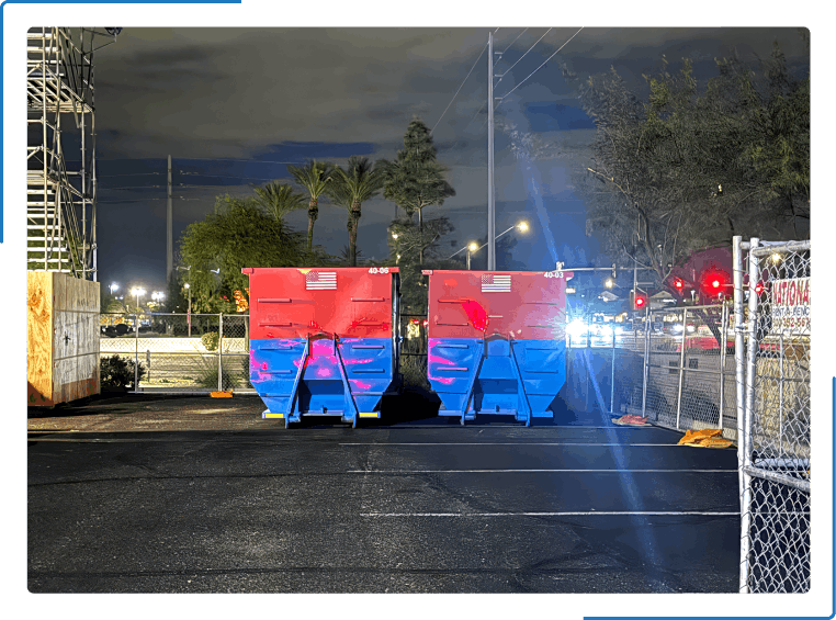 Two green dumpsters in a parking lot at night with lights and a fence in view.