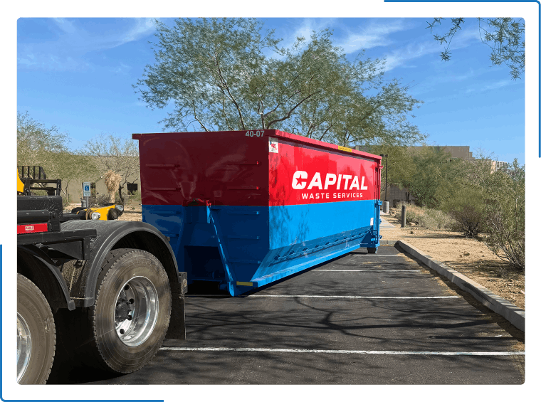 A red and blue Capital Waste dumpster sits on an asphalt residential street under a blue sky.