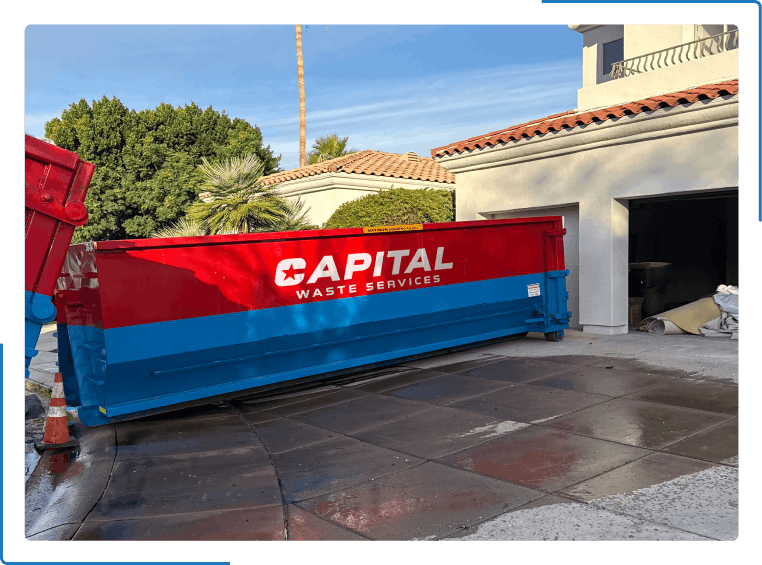 A red and blue Capital Waste Services dumpster sits in a driveway in front of a residential garage.
