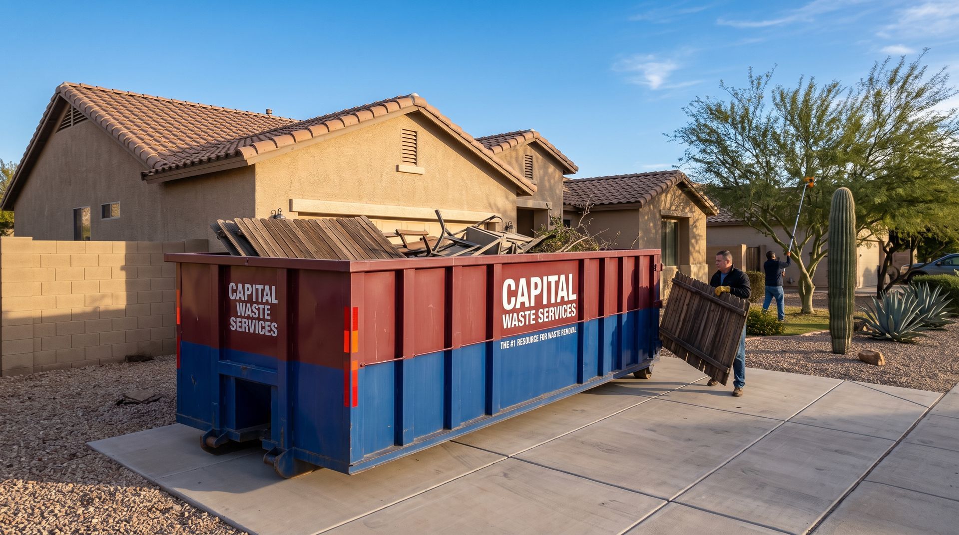 Large red-and-blue dumpster beside a suburban house and sidewalk under a clear blue sky