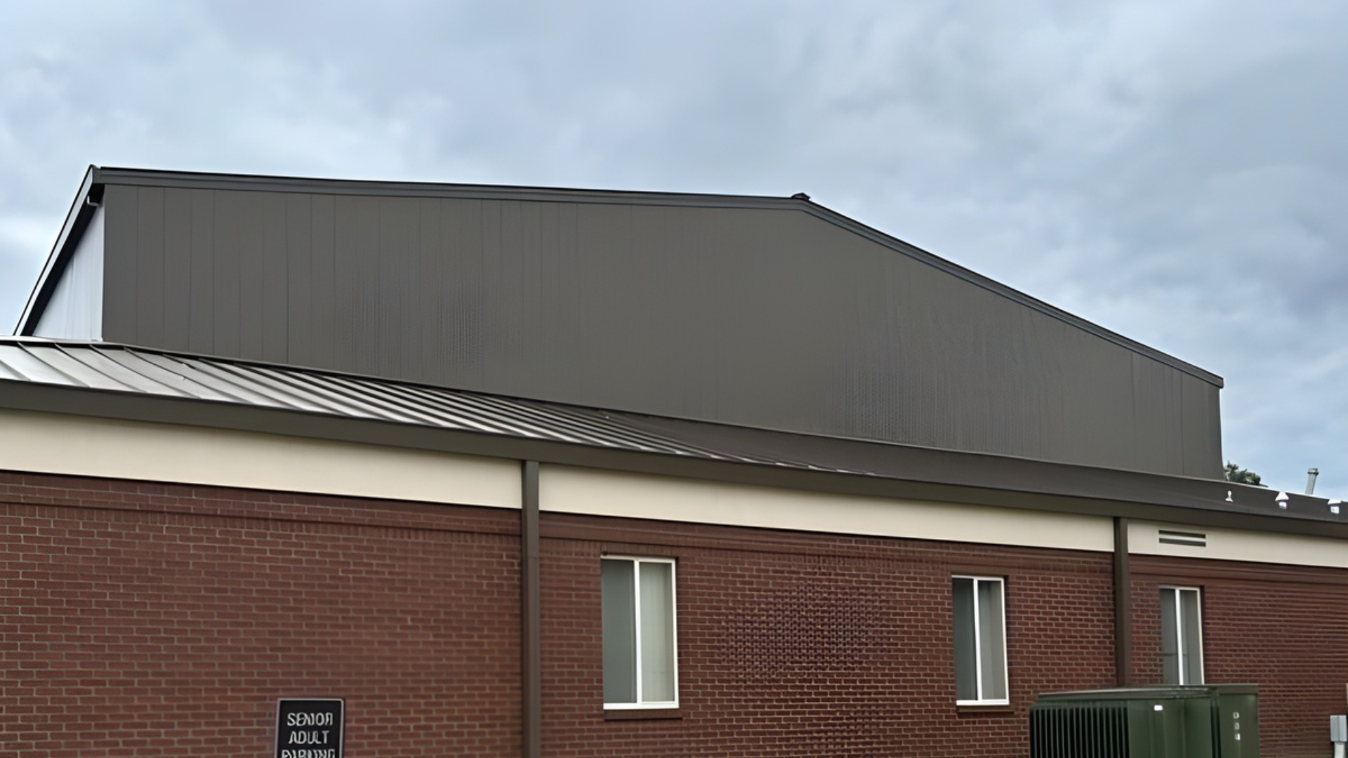 Building exterior with brown brick, a light-colored top, and a large, gray, angled roof against a cloudy sky.