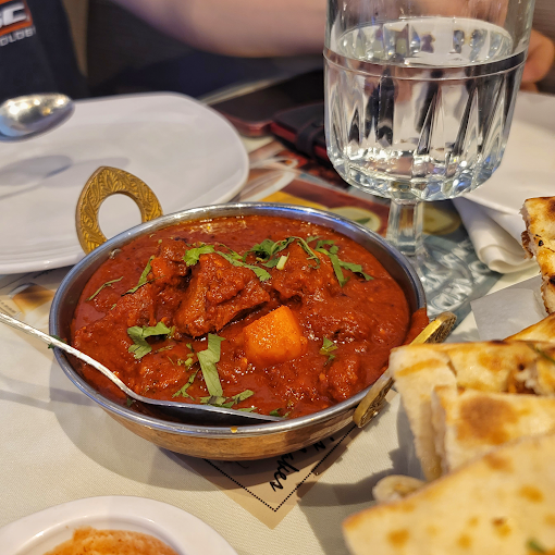 A bowl of red curry with meat and vegetables, naan bread, and a glass of water on a table.