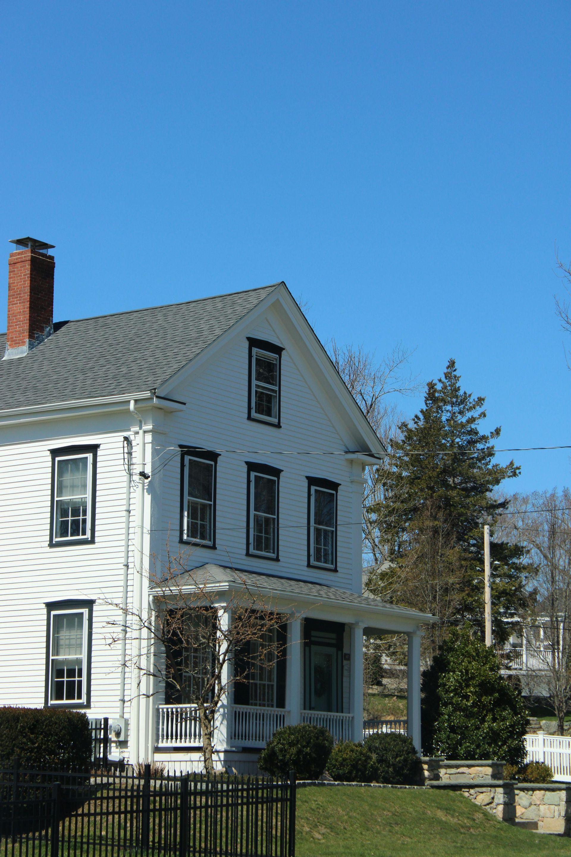 White two-story house with porch, gray roof, and brick chimney under a clear blue sky