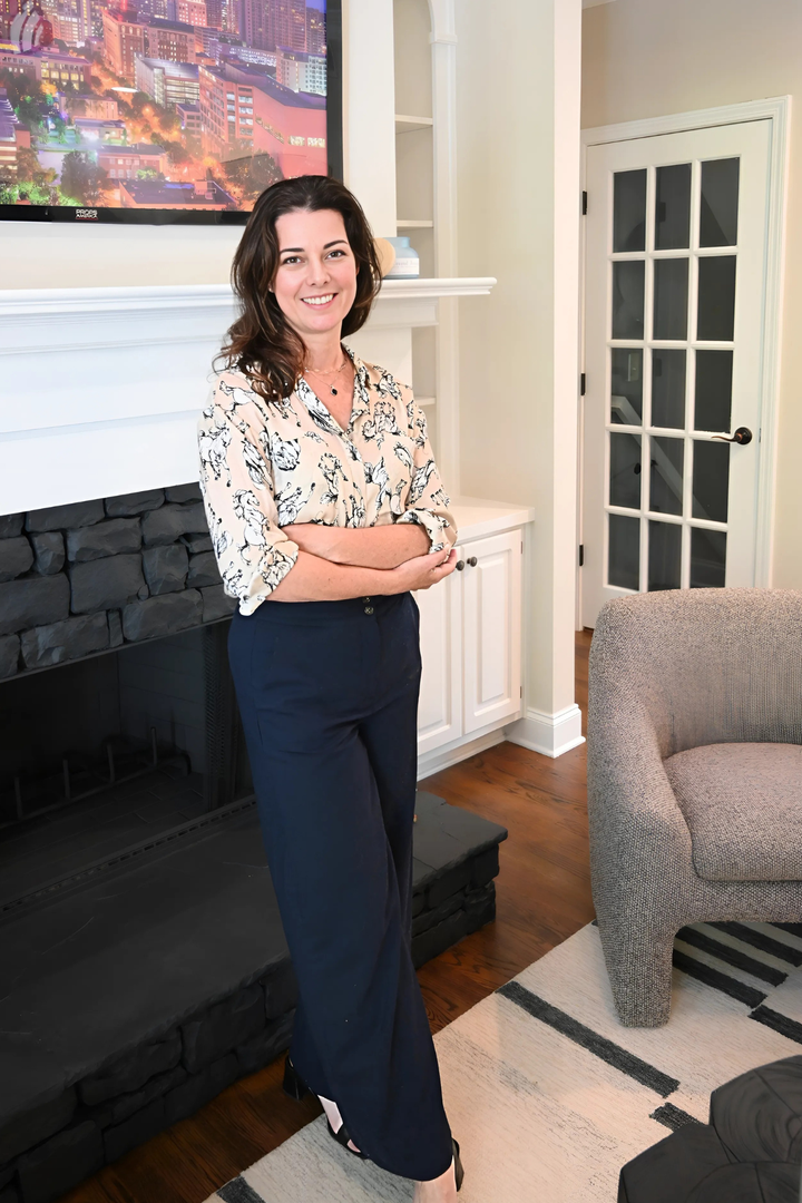 Woman in a floral top and navy pants stands in a living room, arms crossed, smiling.