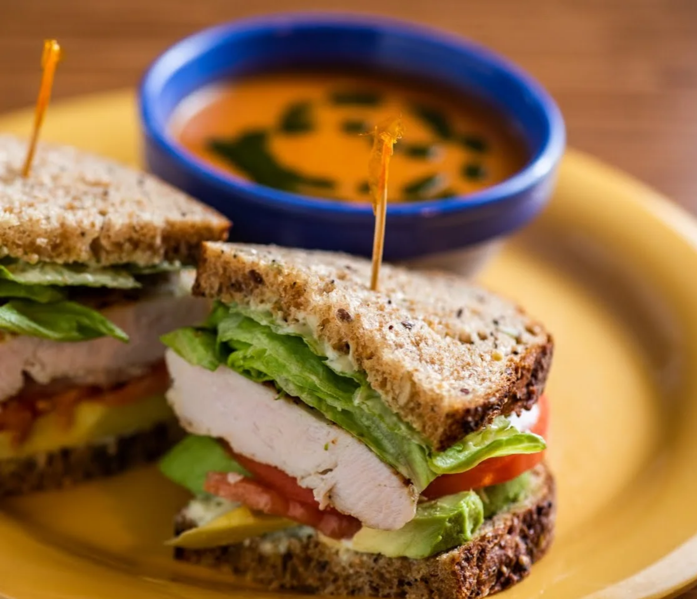 Chicken sandwich with soup, on a yellow plate; brown bread, green lettuce, red tomato, orange soup in a blue bowl.