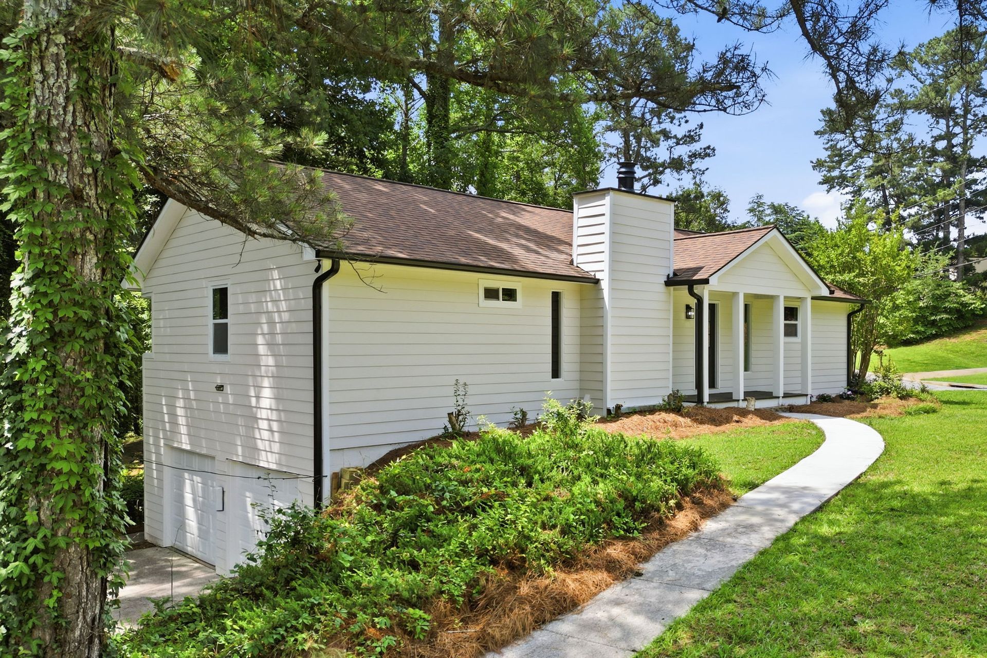 White cottage with brown roof and curved sidewalk, surrounded by green lawn and trees