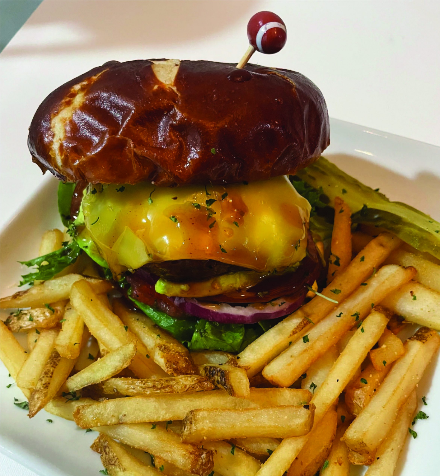 Burger on pretzel bun with fries and pickle on a white plate.