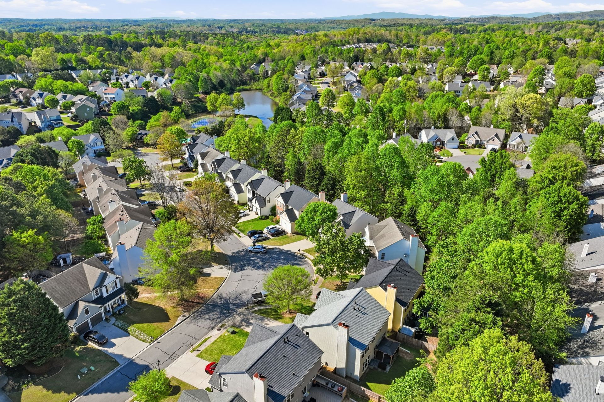 Aerial view of a suburban neighborhood with houses, tree-lined streets, and dense green trees under clear skies