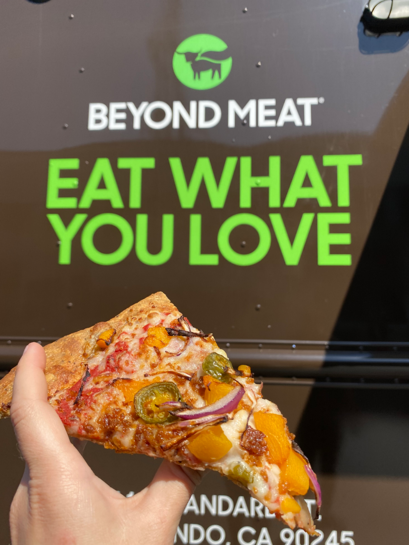 Hand holding slice of pizza with toppings in front of a Beyond Meat food truck.