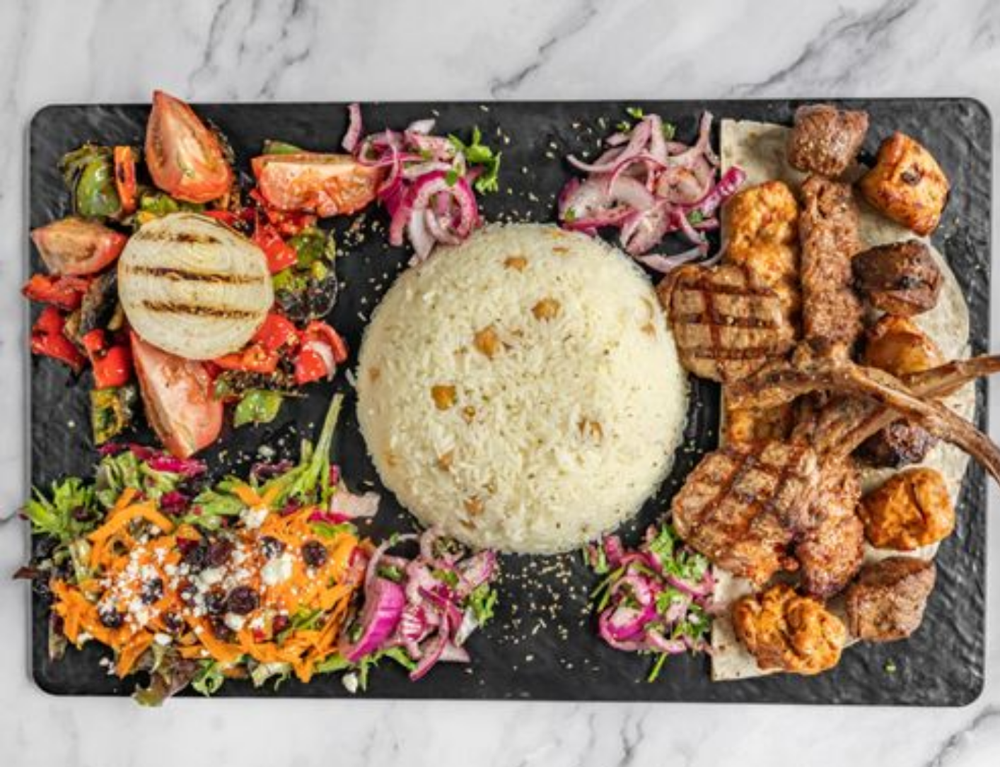 Platter of grilled meats, rice, and vegetables on a black serving board.