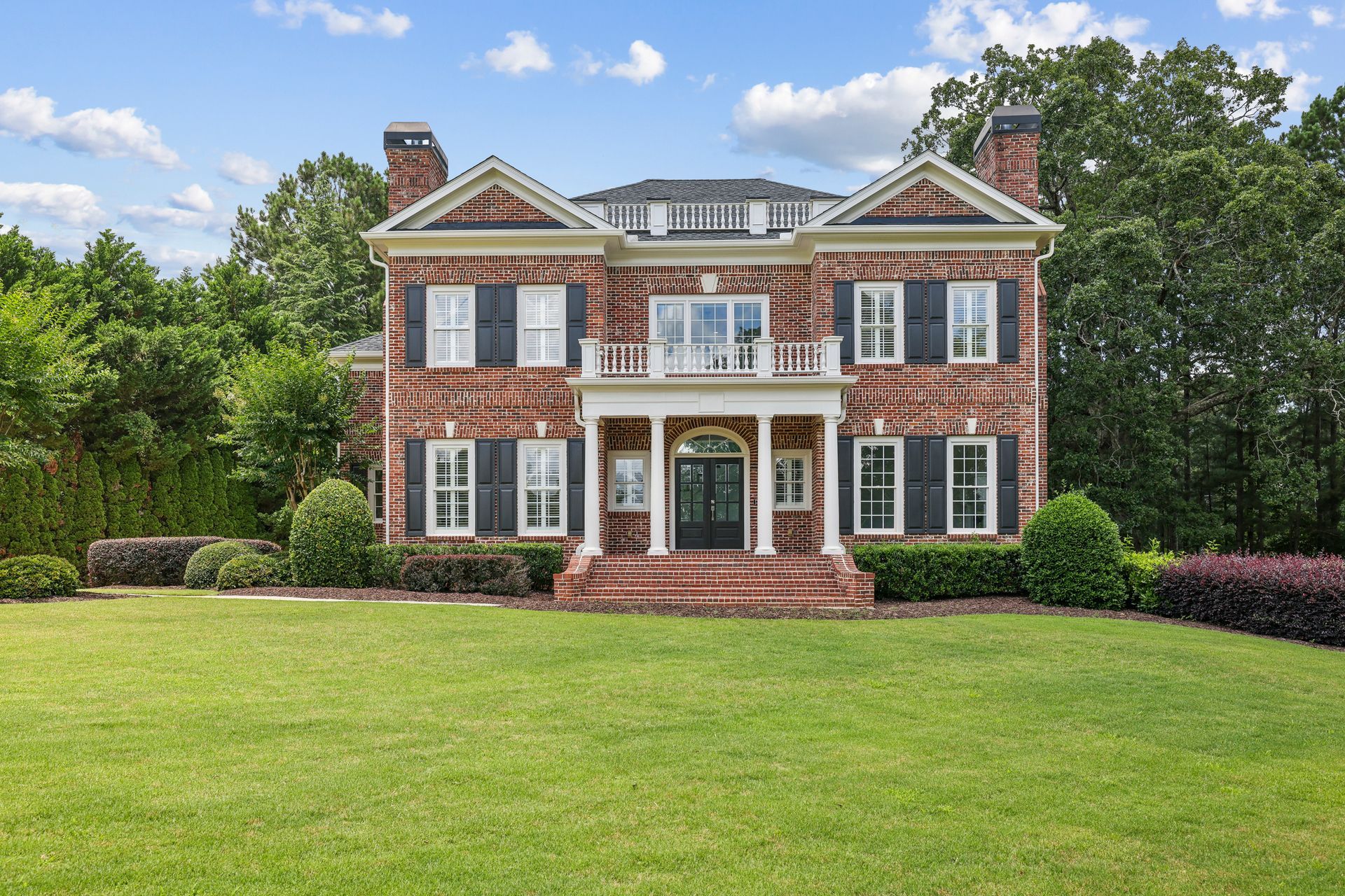 Two-story brick house with black shutters, white columns, and a large front lawn under a blue sky.