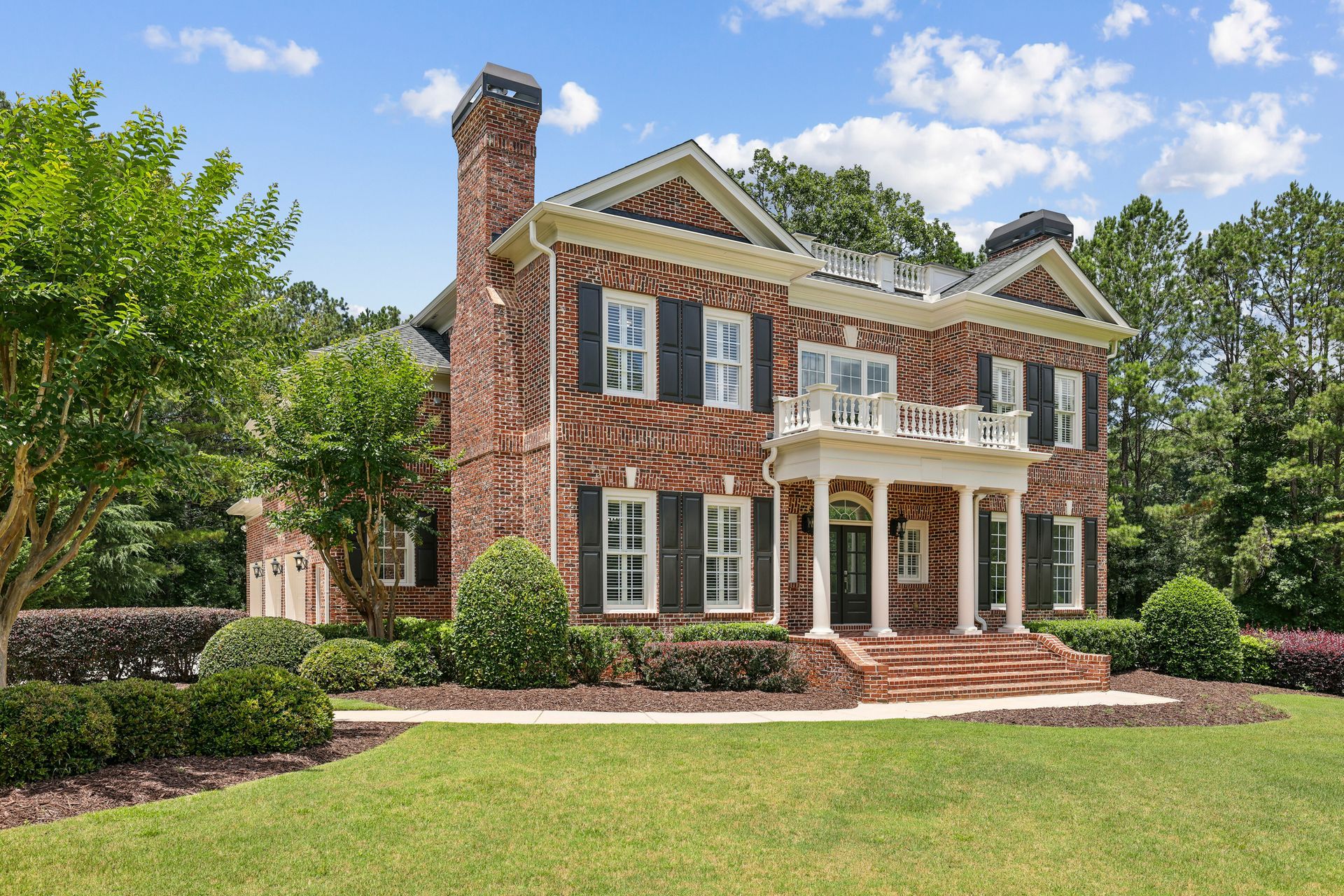 Large brick colonial-style house with white trim, black shutters, and a manicured lawn under a blue sky.