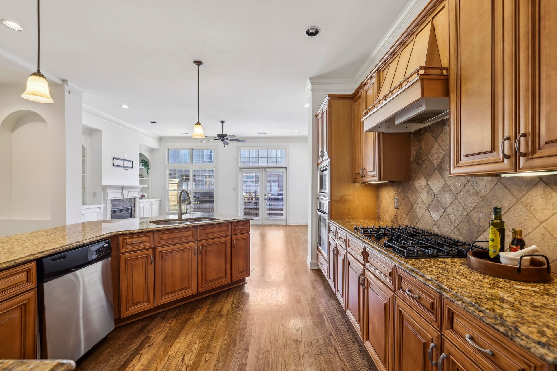 Bright kitchen with wooden cabinets, granite counters, stainless dishwasher, and a hallway to glass double doors.