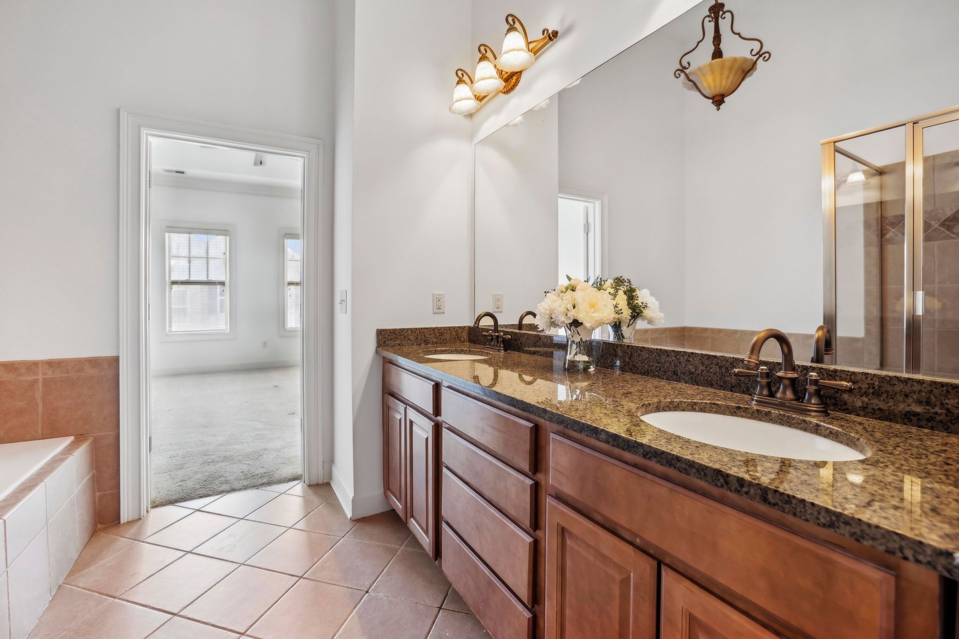 Bright bathroom with granite double vanity, large mirror, tiled floor, and doorway to a sunlit room
