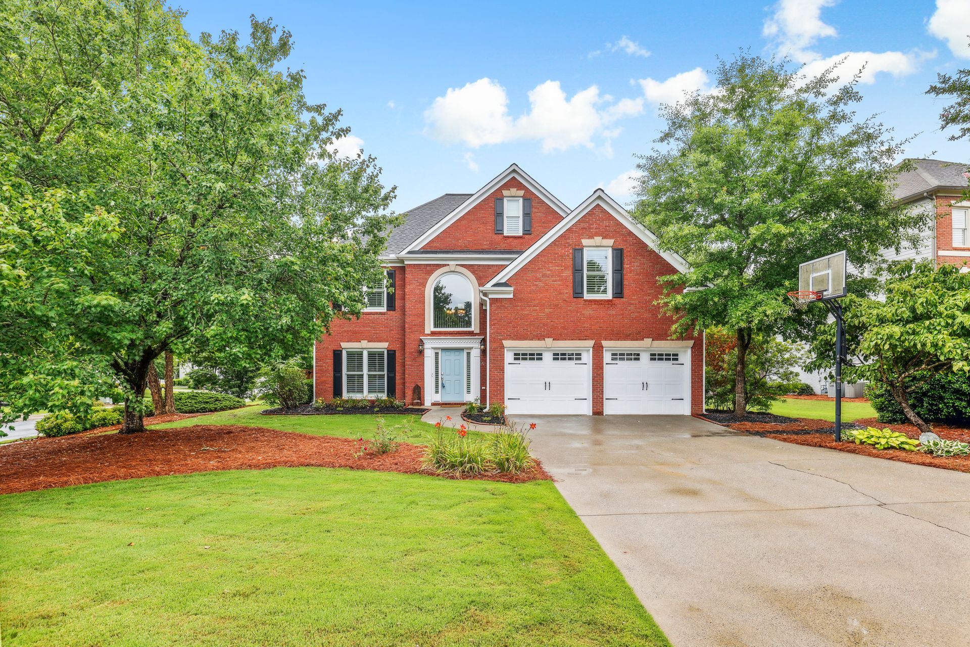 Red brick two-story house with a blue front door, two-car garage, and basketball hoop in the yard.