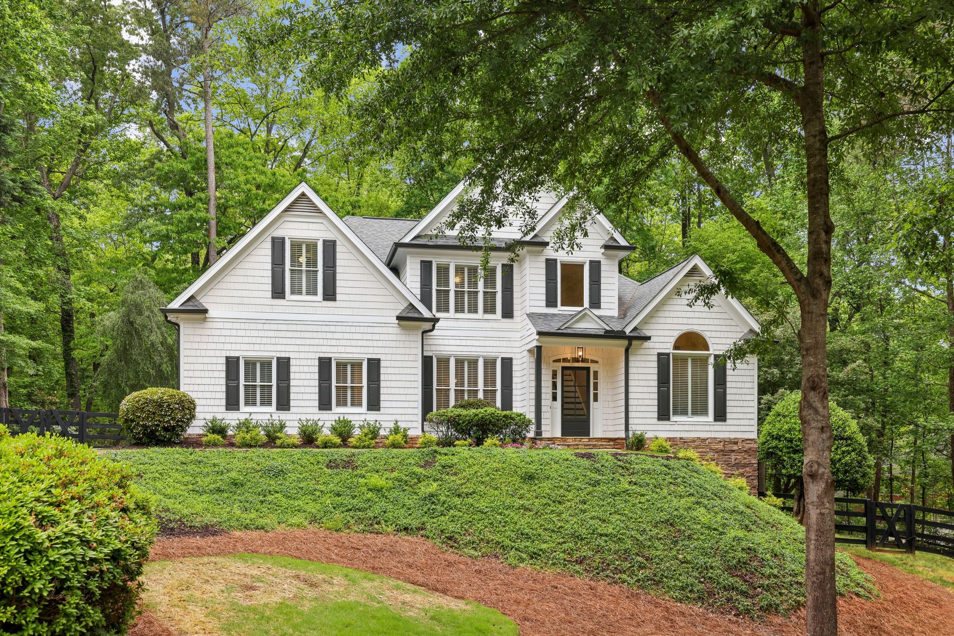 White house with black shutters, on a green, landscaped hillside, surrounded by trees.