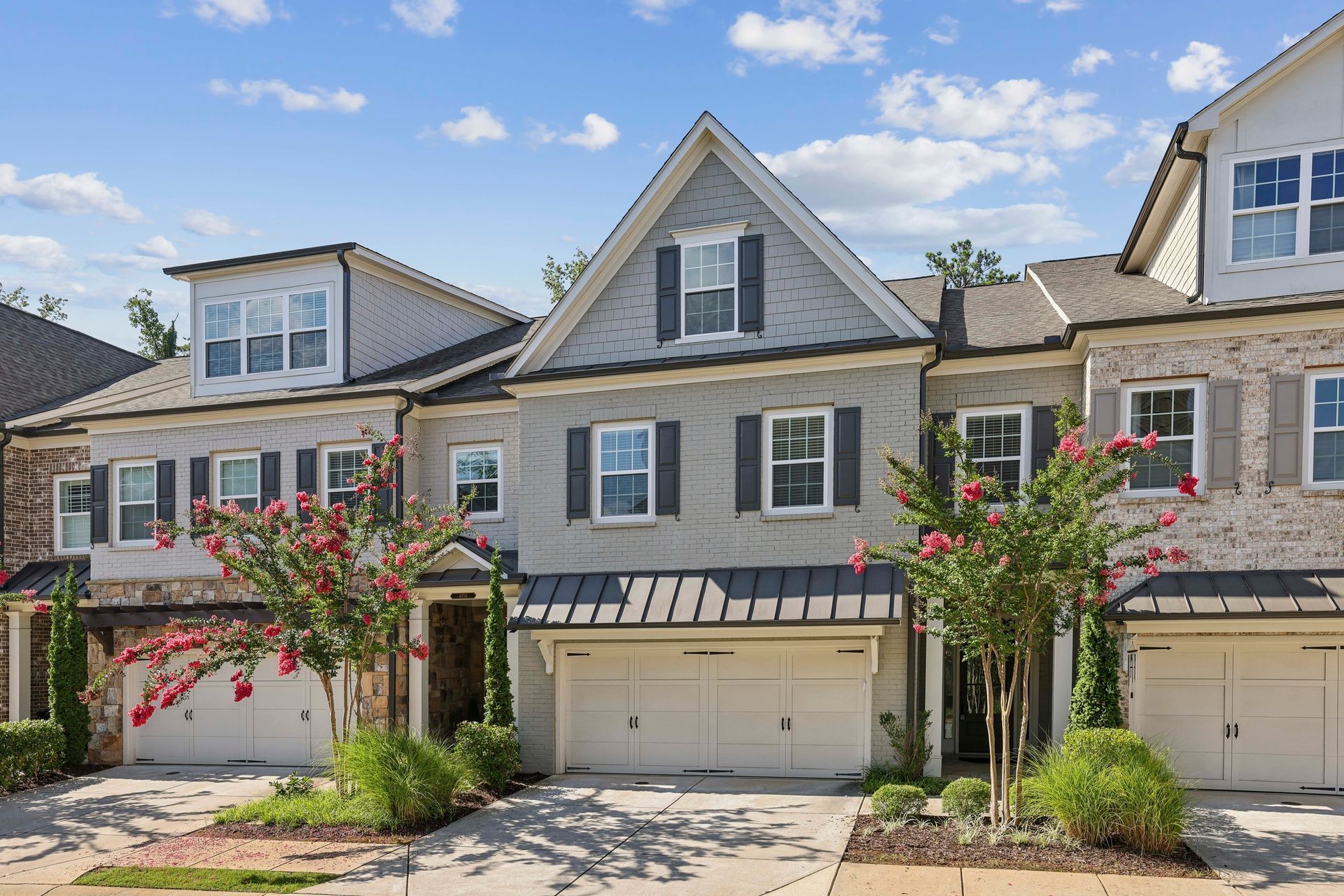 Gray townhomes with white trim, black shutters, and tan garage doors. Flowering trees in front.