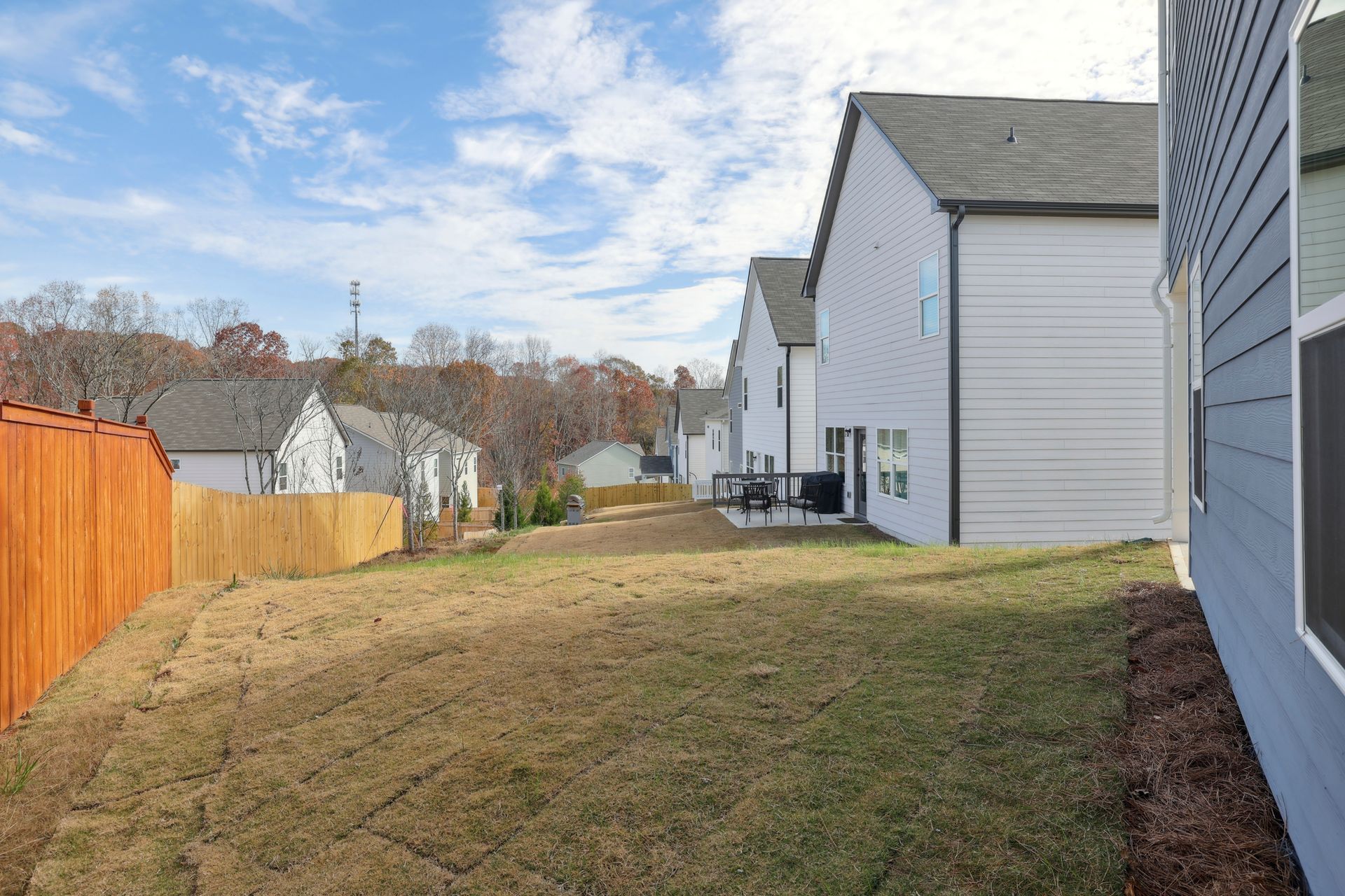 Narrow grassy side yard between white houses and a wooden fence under a partly cloudy sky