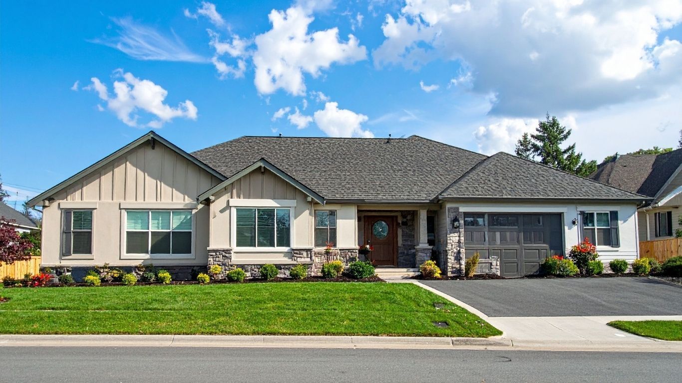 Single-story beige house with a dark shingled roof, front yard, and two-car garage under a bright blue sky.