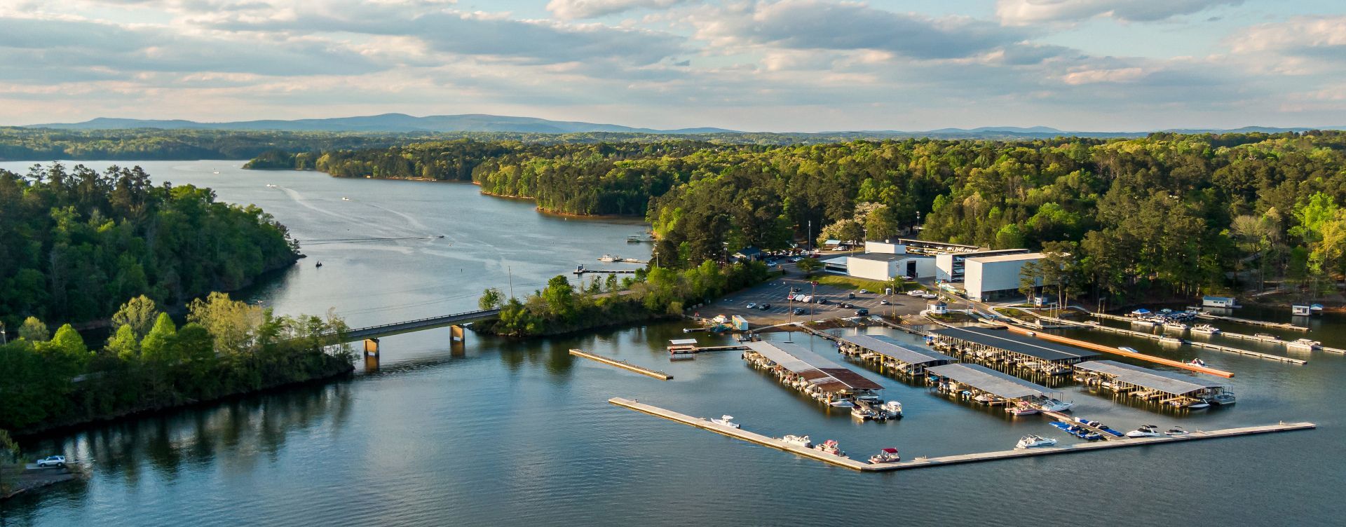 Aerial view of a marina with houseboats, docked on a lake, surrounded by green trees under a cloudy sky.
