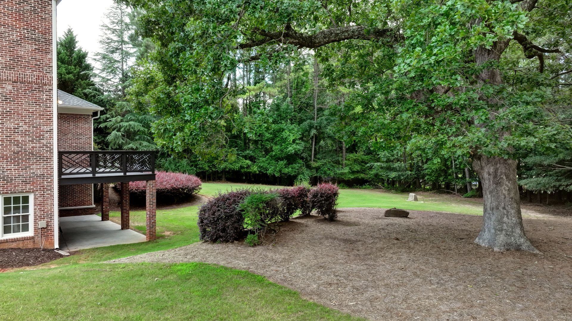 Backyard with brick house, wooden deck, large trees, shrubs, and grassy lawn