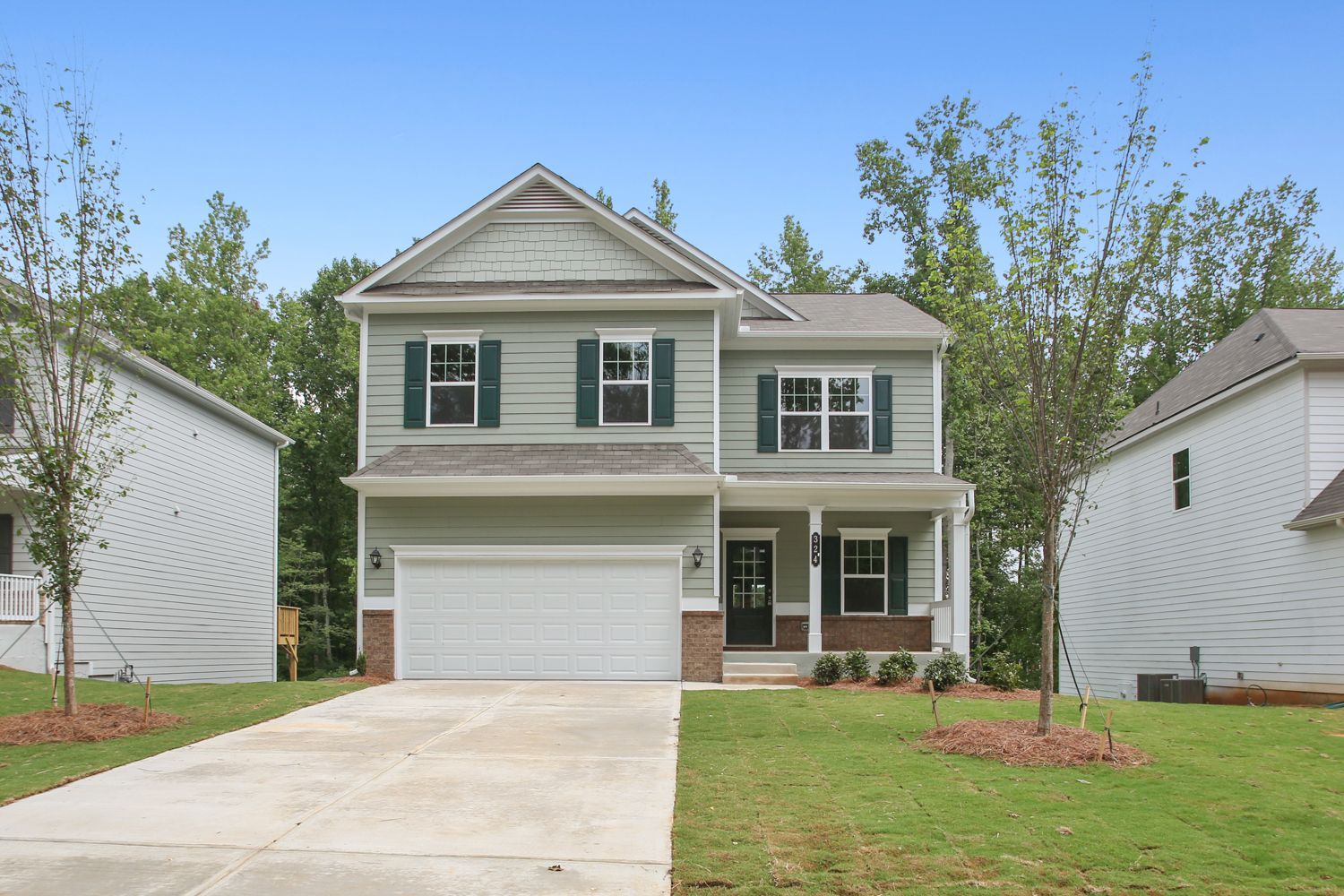 Two-story house with green siding, a white garage door, and a concrete driveway, flanked by other houses and trees.