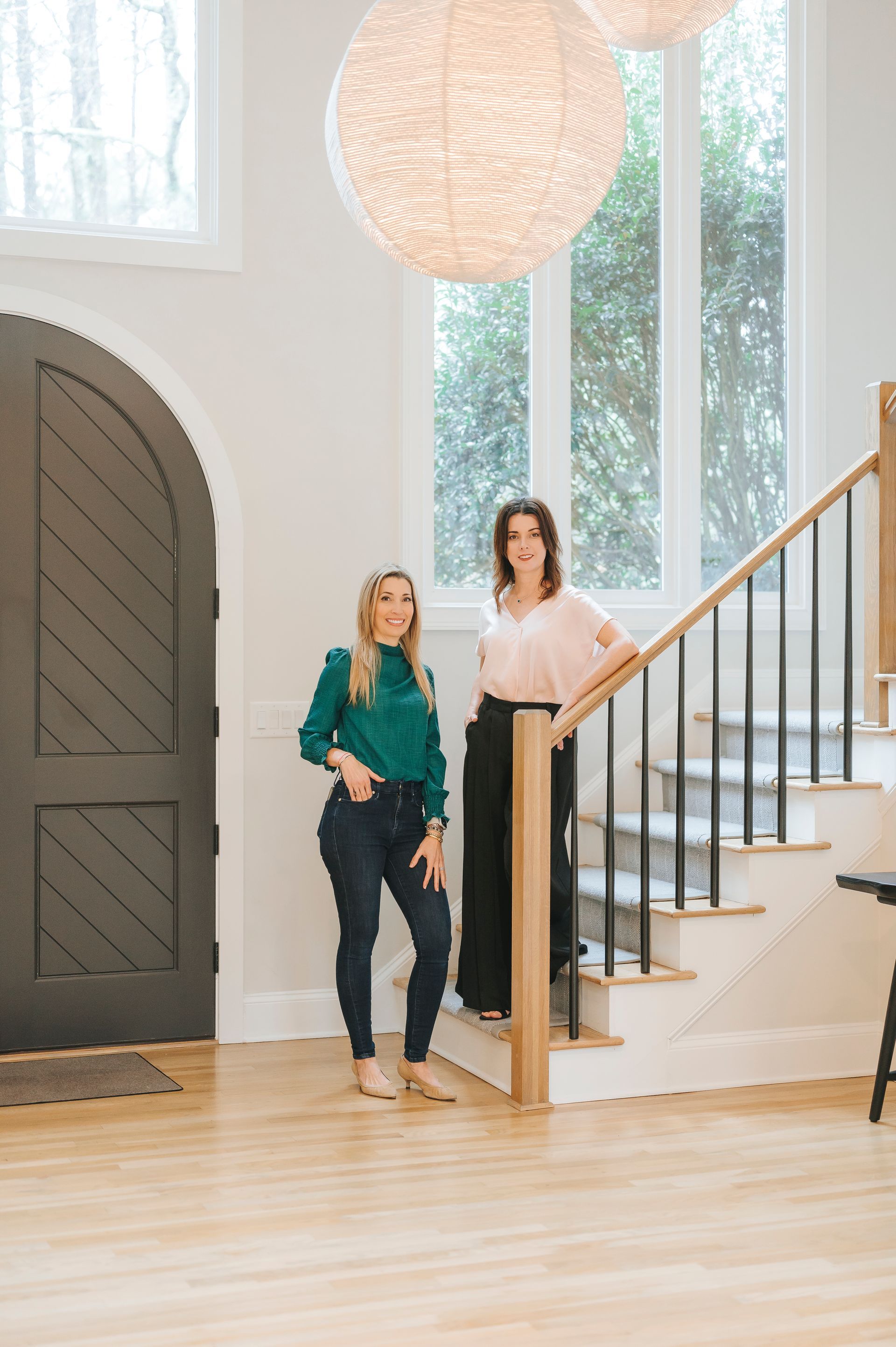 Two women posing in a home's entryway: one in green, jeans; the other in pink, black pants.