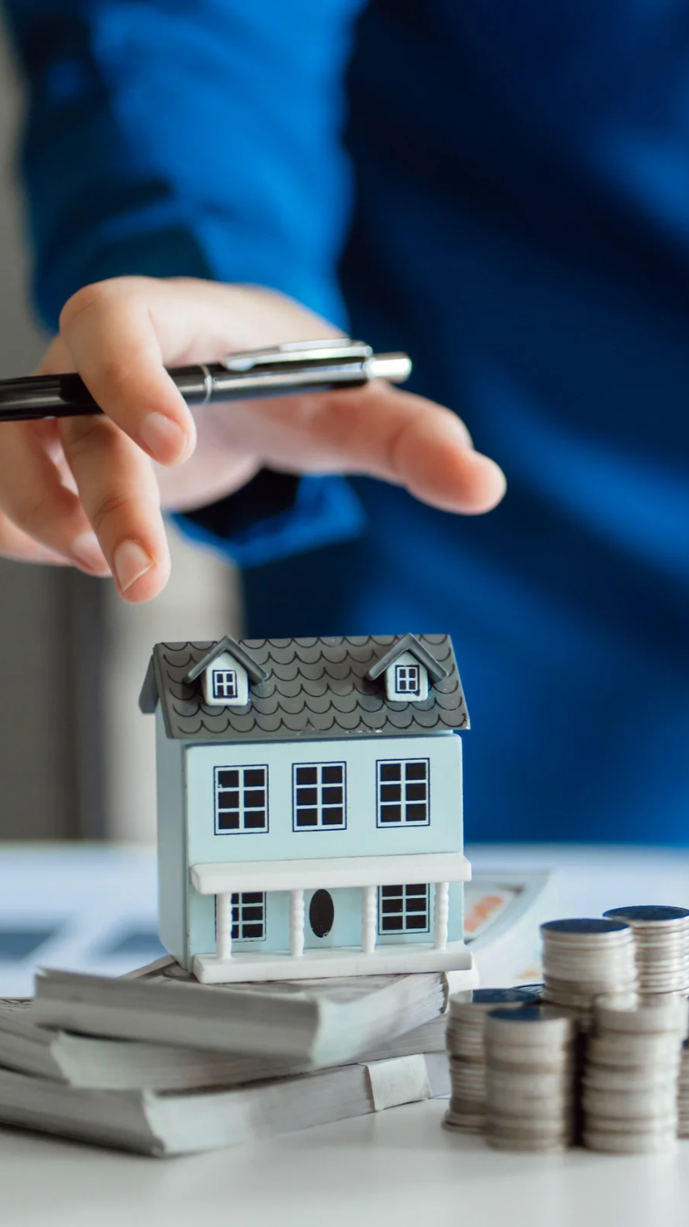 Hand hovering over a small house model beside stacked coins and paperwork, suggesting home finance or mortgage planning
