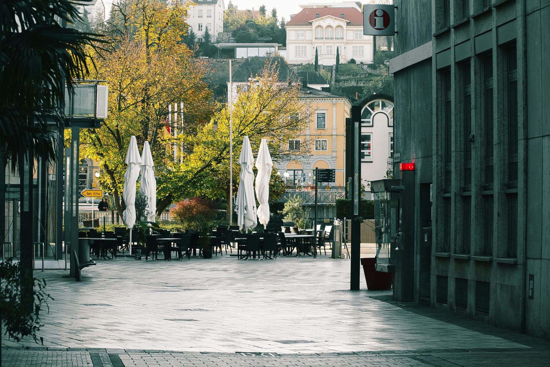 A sunny town square with tables, white umbrellas, and large trees in front of traditional buildings.
