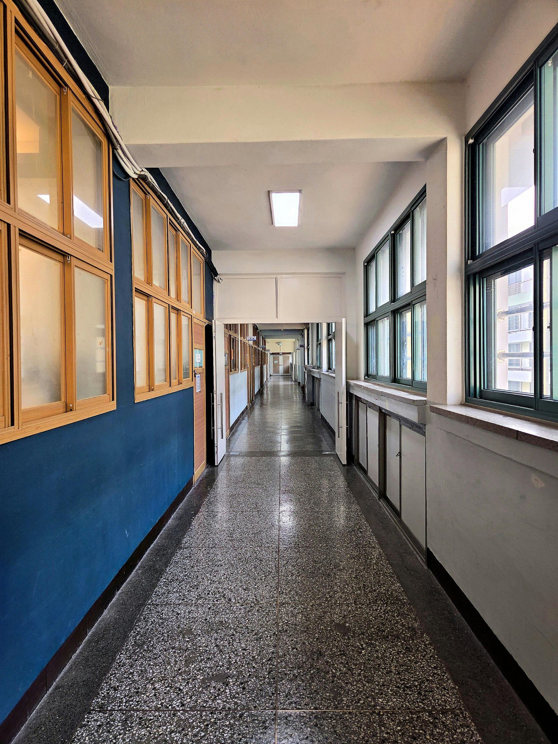 A long, empty indoor hallway with dark blue walls on the left, windows on the right, and a speckled terrazzo floor.