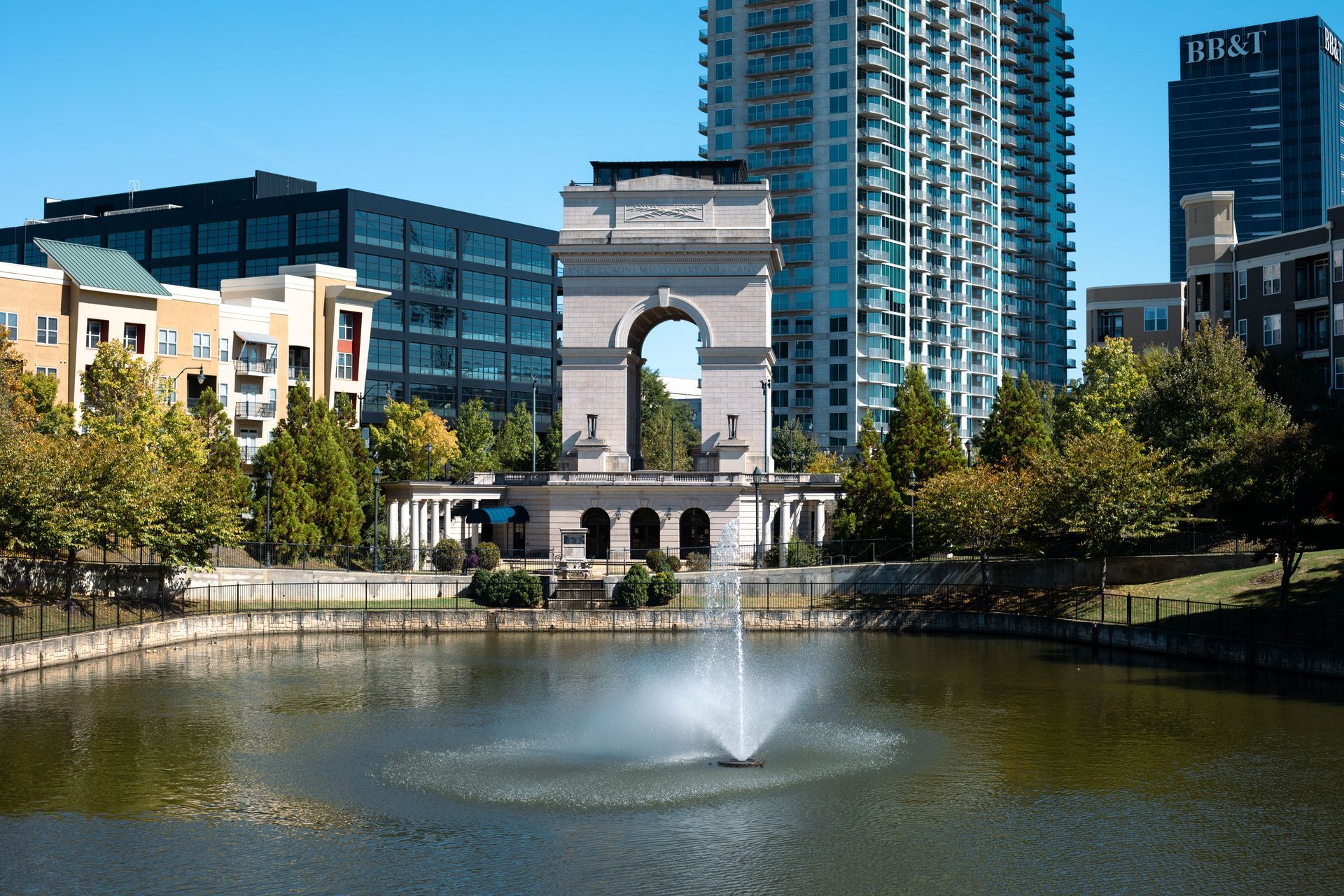 Urban park pond with fountain and a stone arch monument, surrounded by modern city buildings.