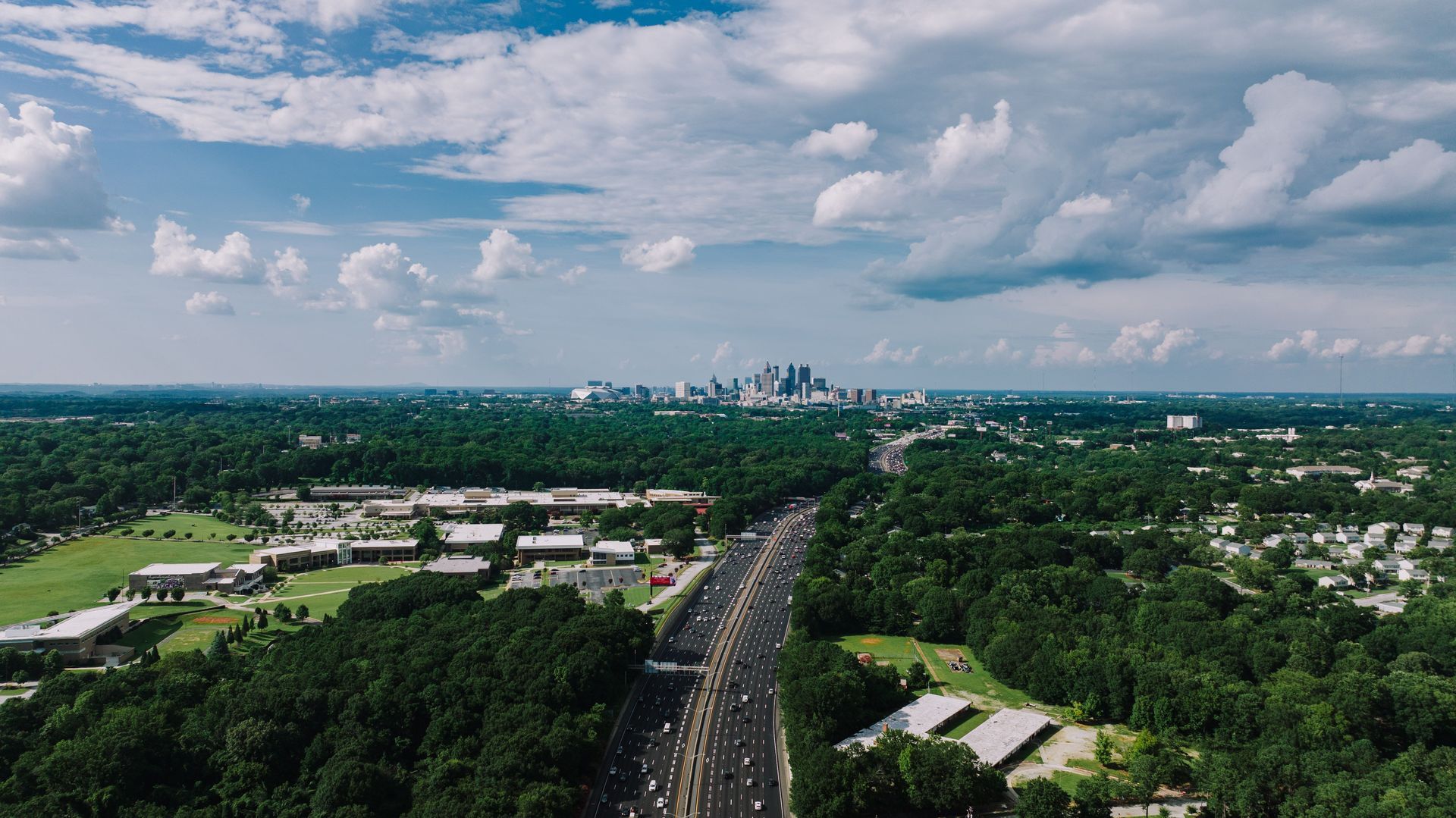 Aerial view of a tree-lined highway leading toward a distant city skyline under a partly cloudy sky