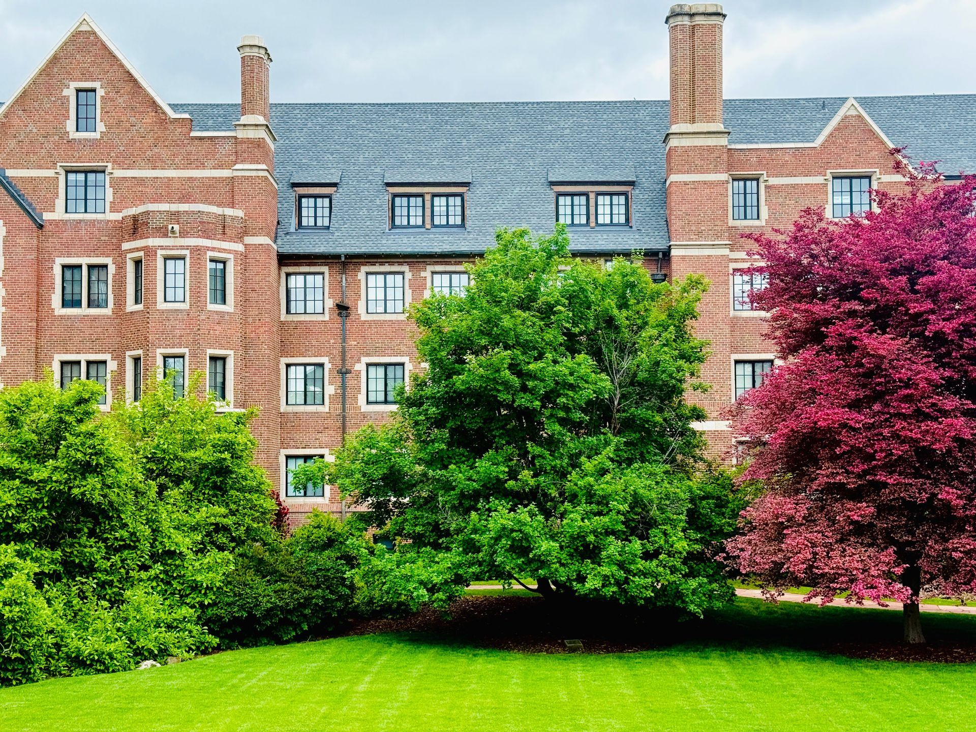 Brick academic building behind green lawn and trees, with a pink tree on the right and a gray roof.