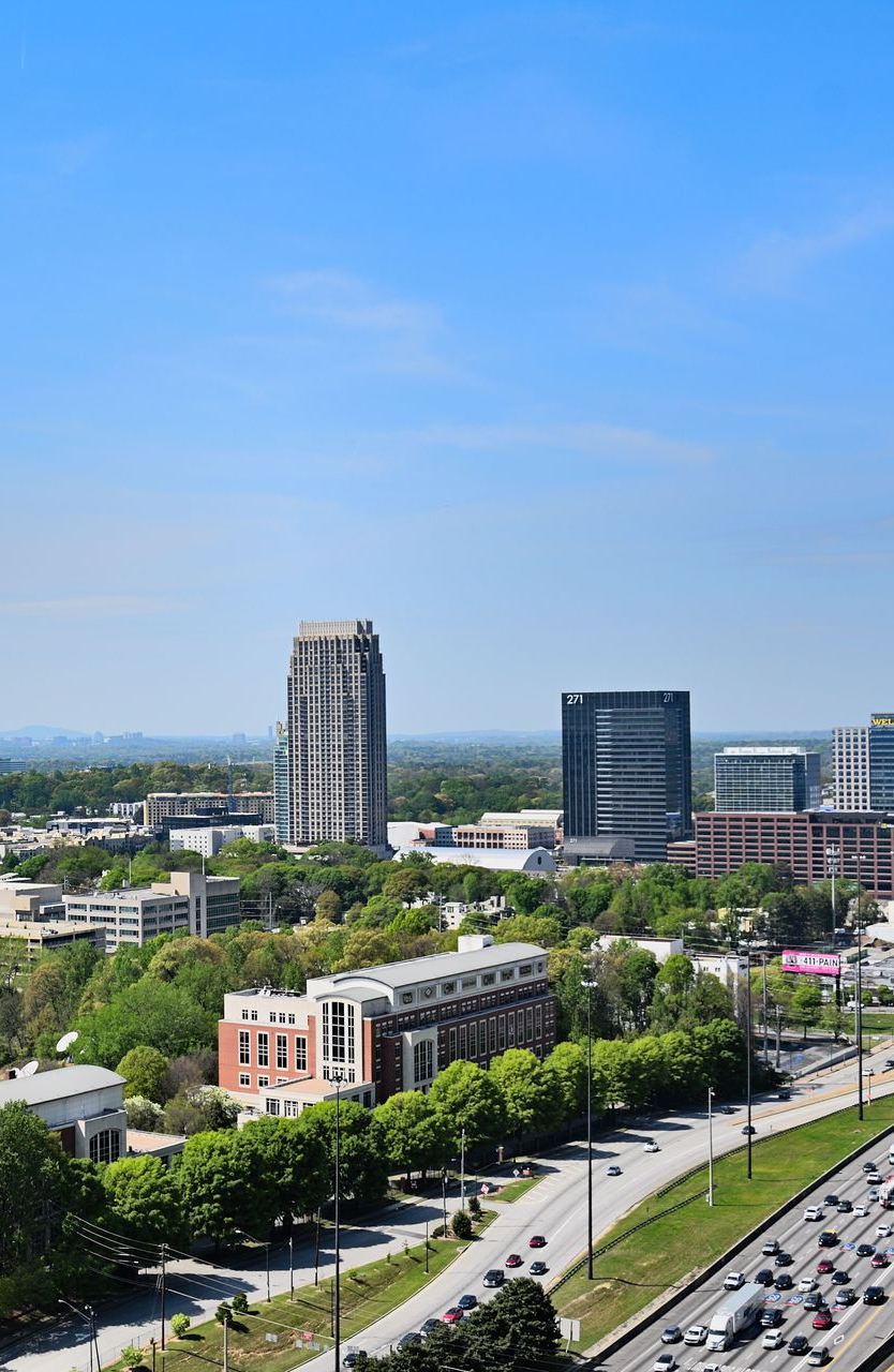 City skyline with tall buildings, tree-lined roads, and traffic under a clear blue sky.