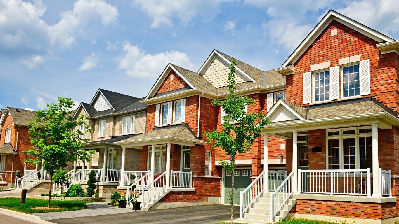 A row of modern brick suburban townhouses with gabled roofs, white porches, and front lawns under a blue sky.