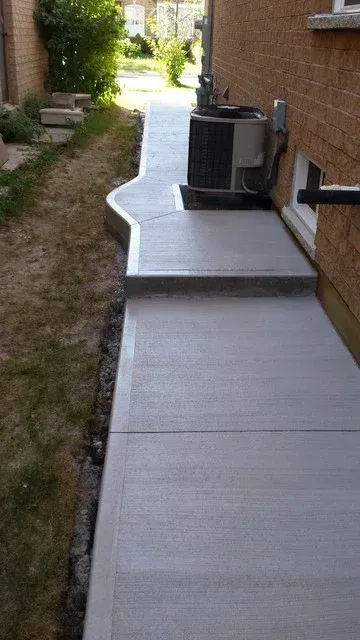 A concrete walkway leading to a house with an air conditioner on the side of it.