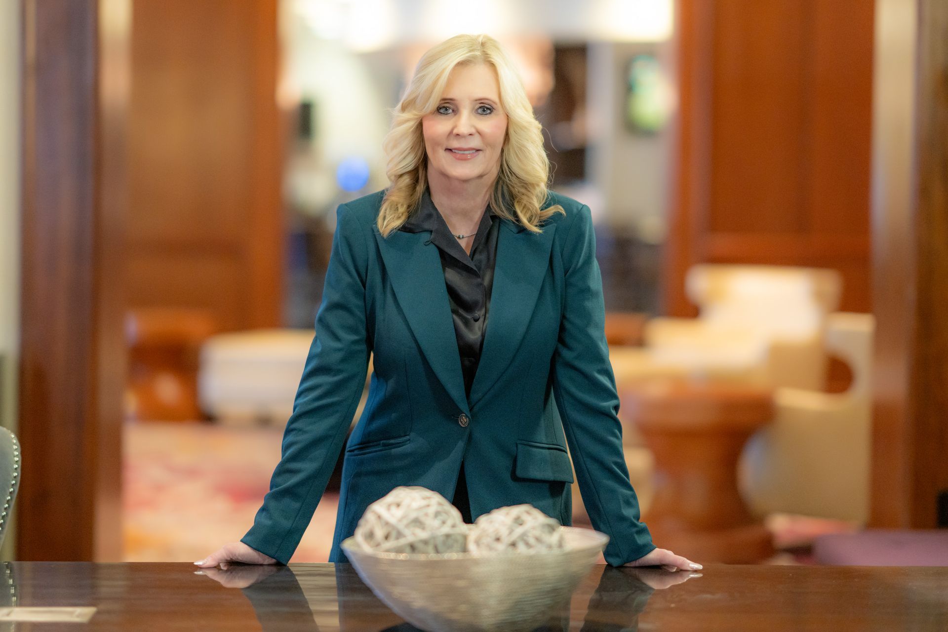 Blonde woman in teal blazer stands at wooden table, arms resting. Formal setting, neutral background.
