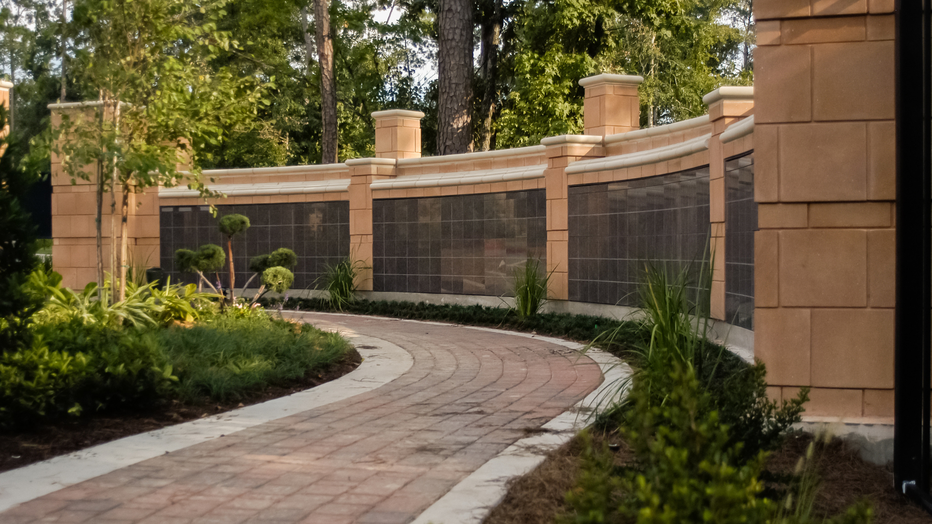 Brick pathway curves past a tan stone columbarium with bronze-toned niches, set in a garden.