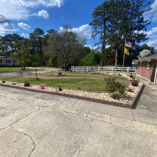 A brick-lined garden with a lawn, small trees, and a white fence in front of a building. Blue sky background.