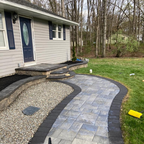 Paved walkway leading to a house with steps and a retaining wall, surrounded by gravel, grass, and trees.