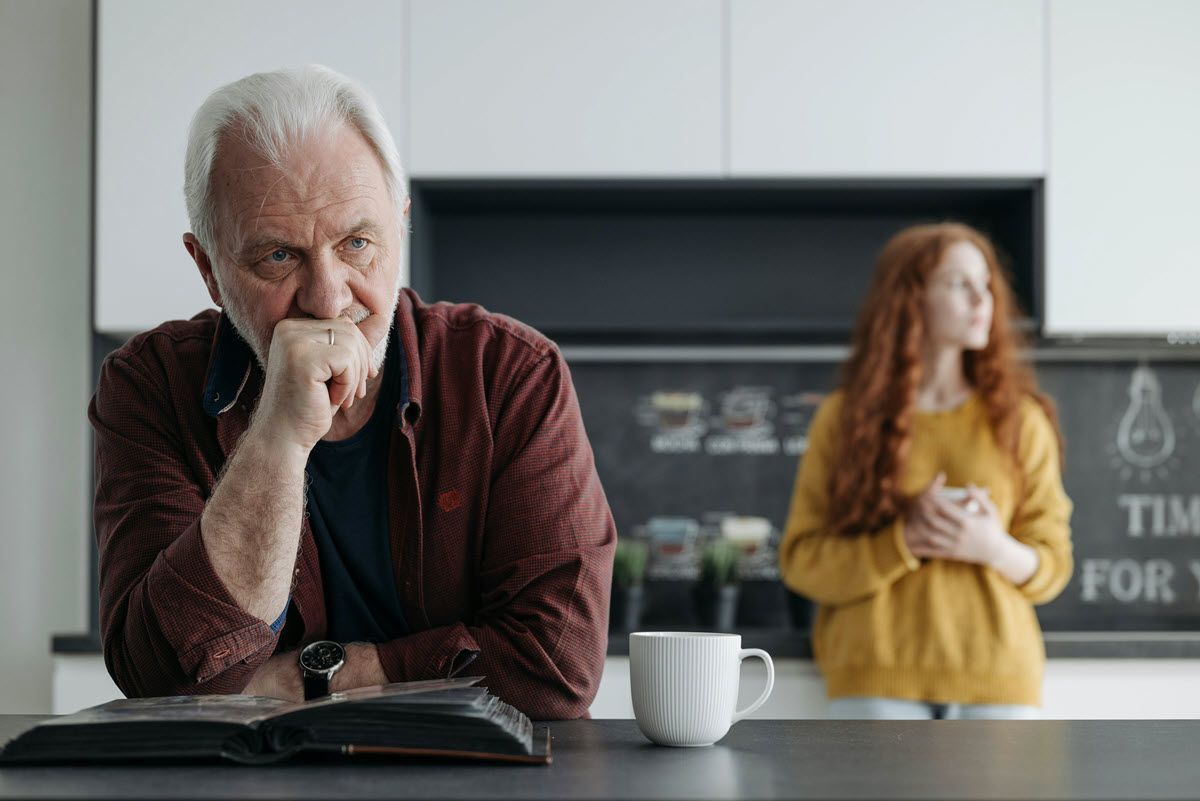 Thoughtful man sitting at a kitchen island, with a woman blurred in the background near a coffee mug.
