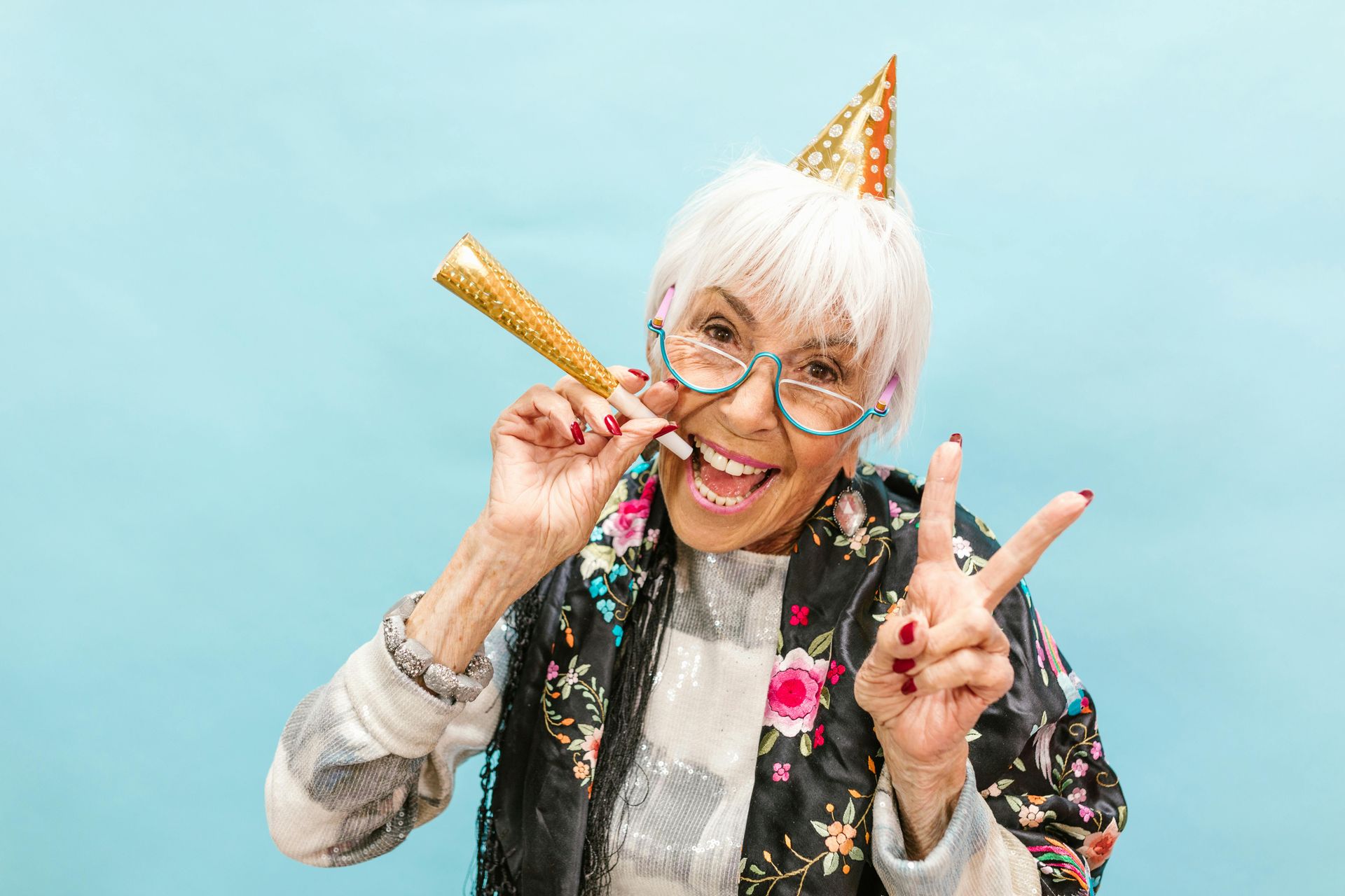White-haired woman with party hat blowing noisemaker, holding up peace sign, smiles.
