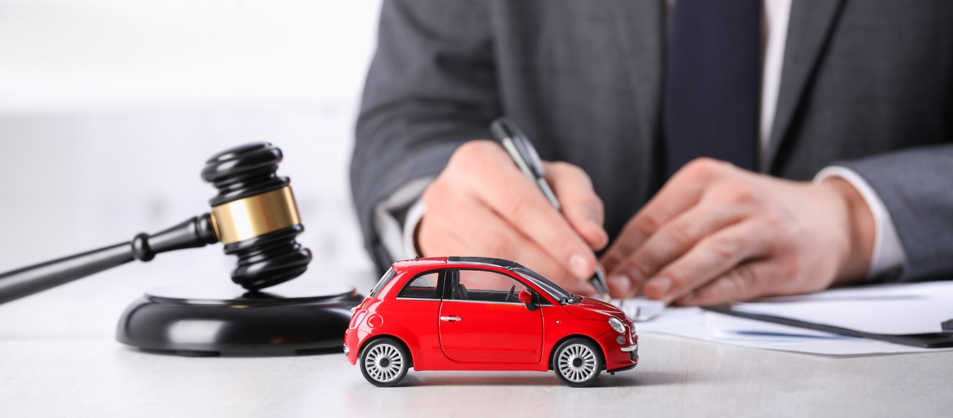 A person in a suit signing paperwork with a red toy car and gavel on the table. A person in a suit signing paperwork with a red toy car and gavel on the table.