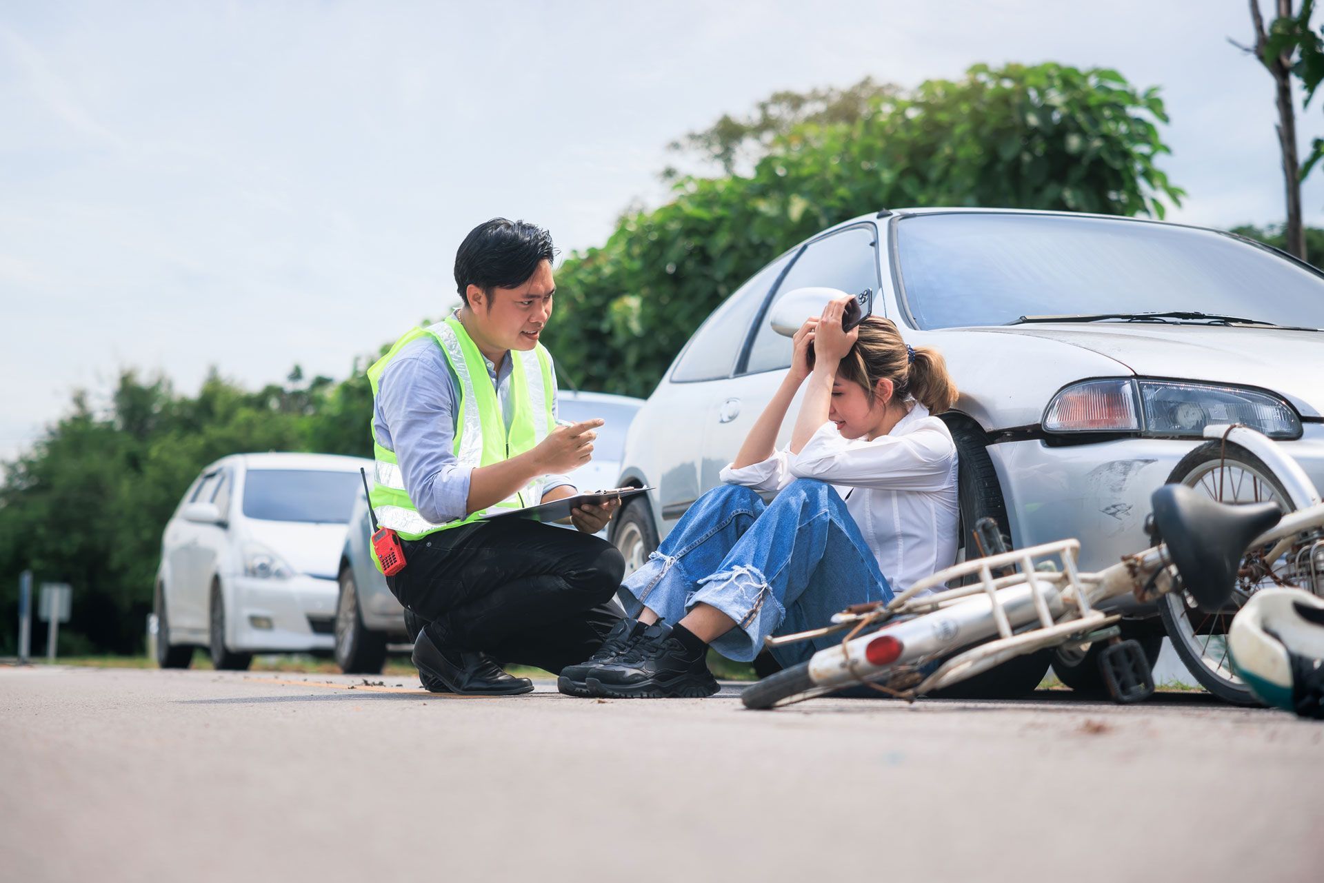 A man in a safety vest is helping an injured woman sitting near a car and bicycle after an accident.