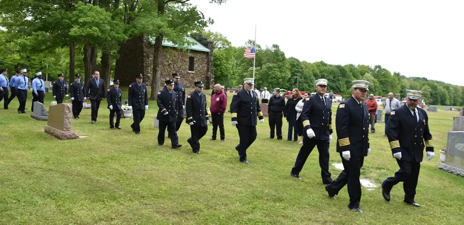 Firefighters march in formation through a cemetery with headstones, toward a small stone building, American flag visible.